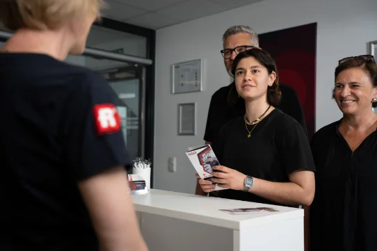 Vier Personen stehen an einem Empfangstresen; eine Frau hält Unterlagen
