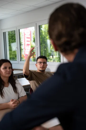 Ein Teilnehmer hebt während einer Besprechung die Hand; zwei weitere Personen sitzen im Raum